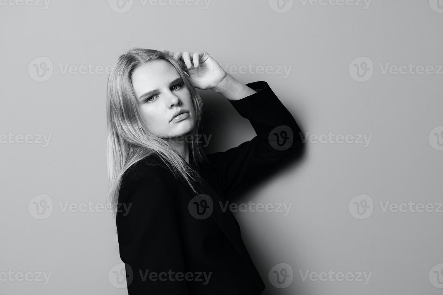 echt jong blond vrouw leunt Aan de studio muur Holding hand- overhead looks Bij camera poseren geïsoleerd in zwart jasje foto