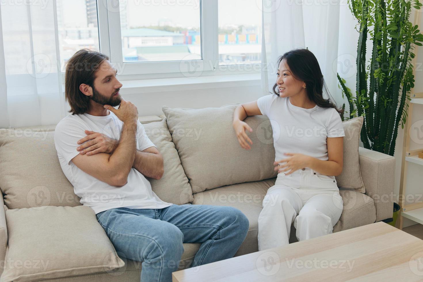 een Mens en een vrouw zittend Bij huis Aan de bankstel in wit elegant t-shirts en chatten vrolijk glimlachen en lachend Bij huis. mannetje en vrouw vriendschap foto