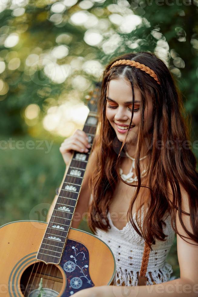 gelukkig hippie vrouw met een gitaar ontspannende in natuur zittend Aan een plaid glimlachen en genieten van de visie. levensstijl in harmonie met natuur en zelf foto