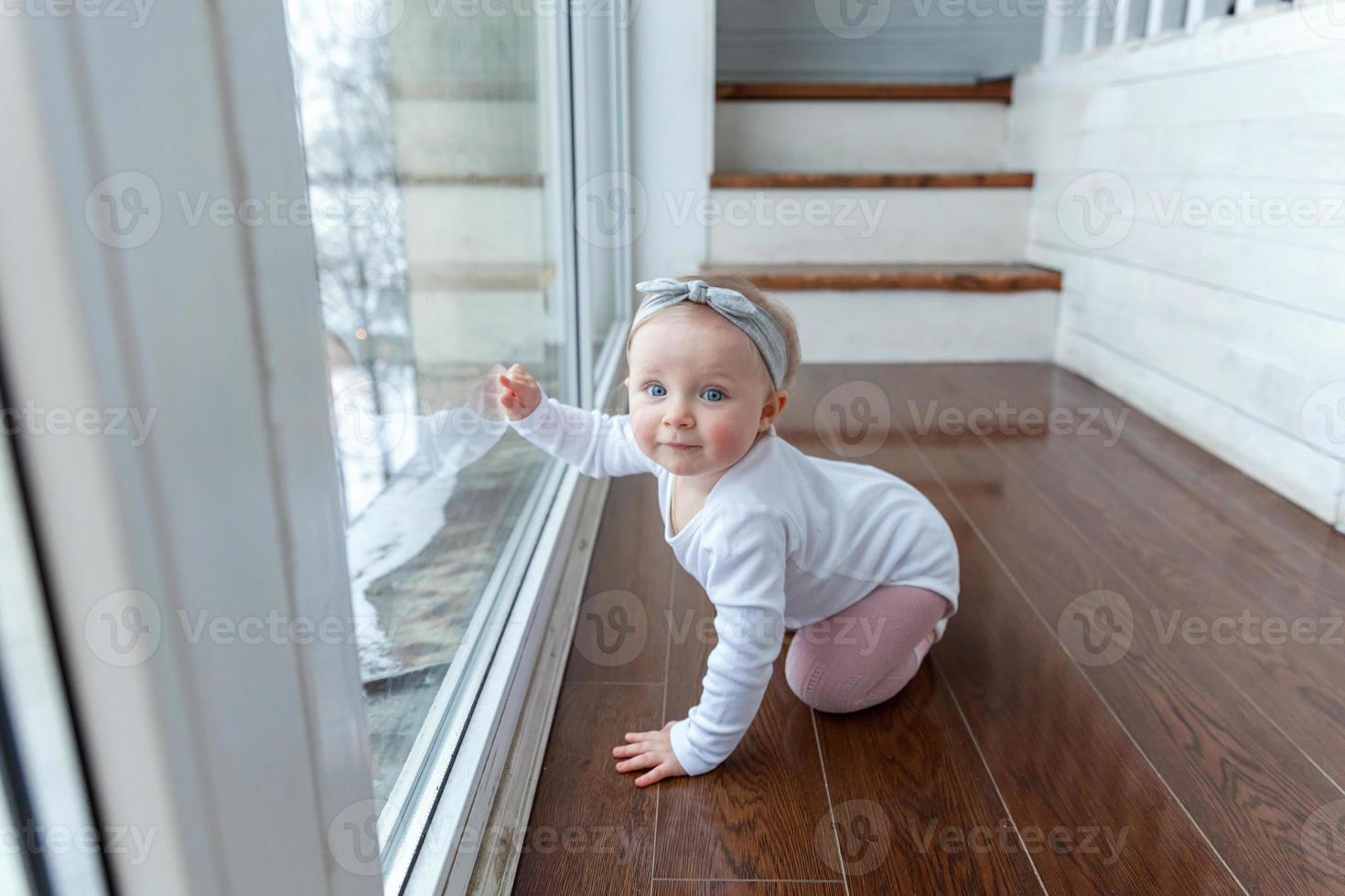 weinig kruipen baby meisje een jaar oud locatie Aan verdieping in helder licht leven kamer in de buurt venster glimlachen en lachend foto