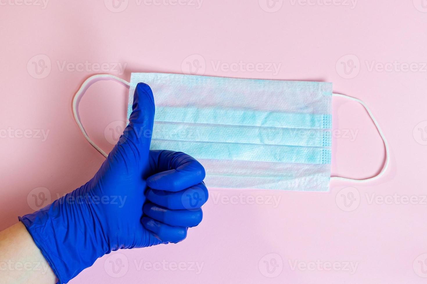een hand in een blauwe latexhandschoen en een chirurgisch masker op een roze achtergrond foto