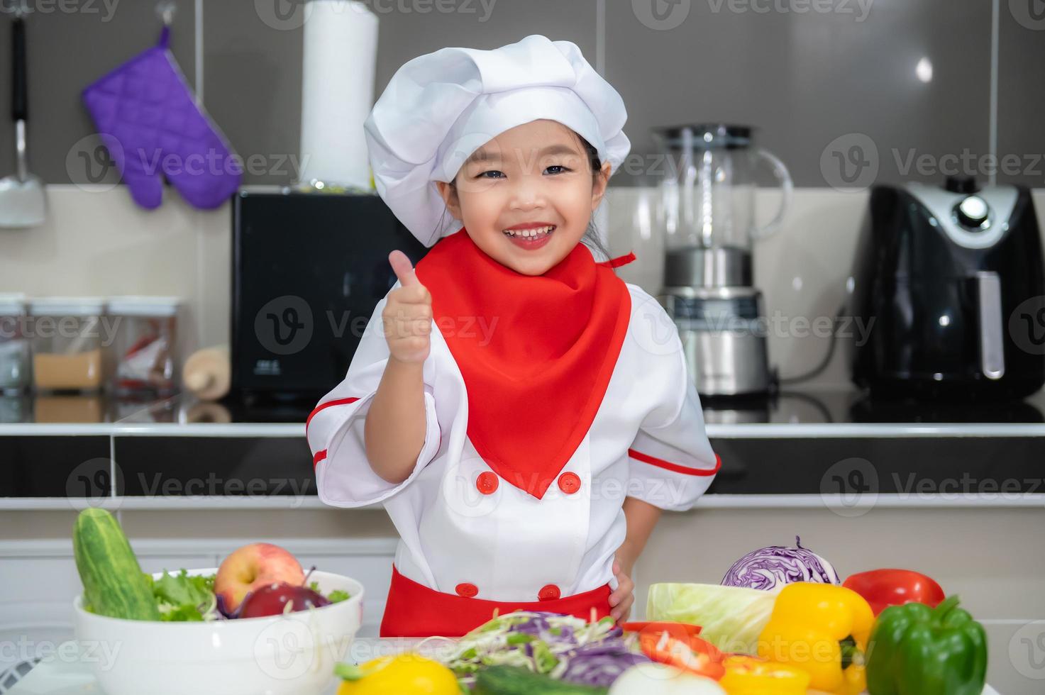 schattig Aziatisch meisje slijtage chef uniform met een veel van vegetarisch Aan de tafel in de keuken kamer, maken voedsel voor eten diner, grappig tijd voor kinderen foto