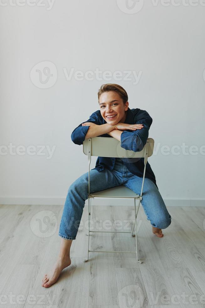 een jong vrouw zittend in een stoel Bij huis glimlachen met tanden met een kort kapsel in jeans en een denim overhemd Aan een wit achtergrond. meisje natuurlijk poses met Nee filters foto