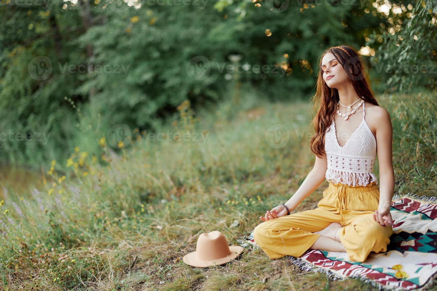 een jong hippie vrouw mediteert in natuur in de park, zittend in een lotus positie Aan haar kleurrijk plaid en genieten van harmonie met de wereld in eco-kleding foto