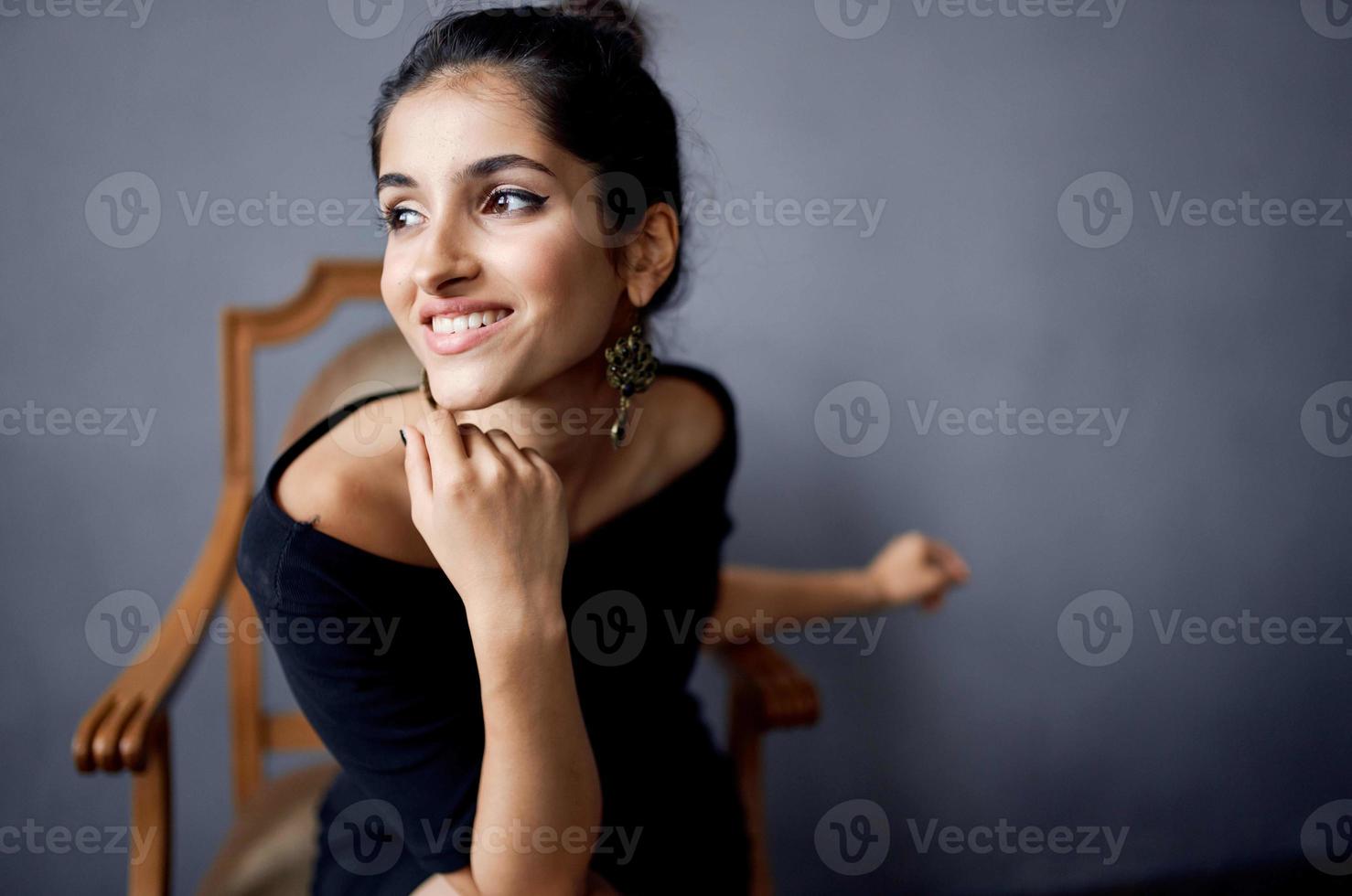 mooi vrouw oorbellen sieraden poseren in de buurt de stoel zwart jurk donker achtergrond foto