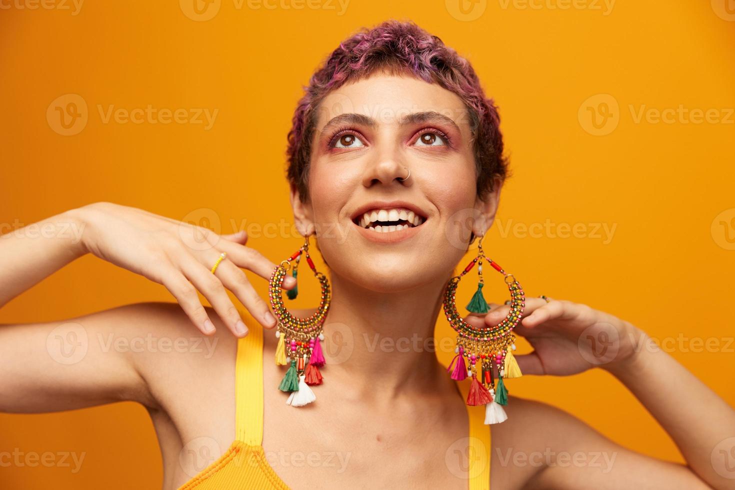 portret van een jong vrouw met een kort kapsel en gekleurde haar- glimlachen en tonen haar tong Bij de camera Aan een oranje achtergrond met oorbellen accessoires in de studio foto