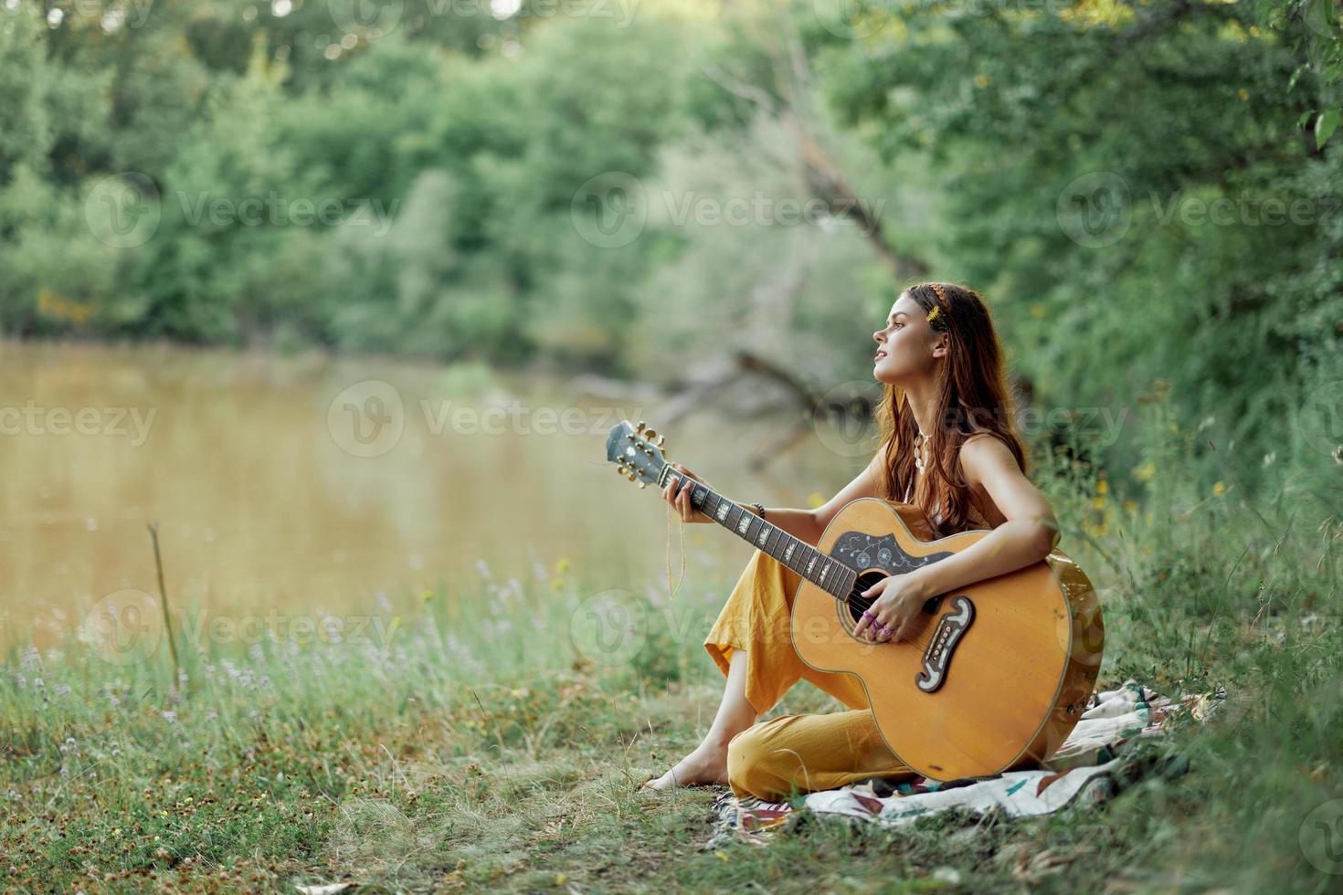 een hippie vrouw spelen haar gitaar glimlacht en zingt liedjes in natuur zittend Aan een plaid in de avond in de zonsondergang zonlicht. een levensstijl in harmonie met de lichaam en natuur foto