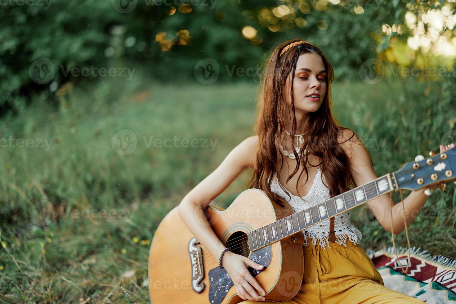 meisje hippie vrouw spelen gitaar in milieuvriendelijk kleren zittend Aan de grond buiten in natuur in de vallen aan het kijken de zonsondergang foto