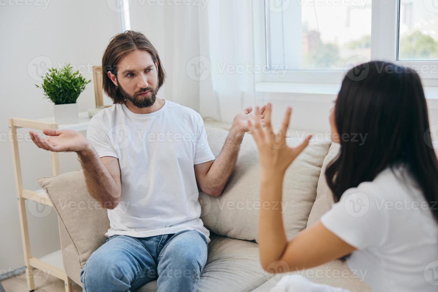 een Mens en een vrouw zitten Aan de bankstel Bij huis in wit t-shirts en chatten schandalig Doen niet begrijpen elk ander. een ruzie in de familie van twee echtgenoten en agressie foto