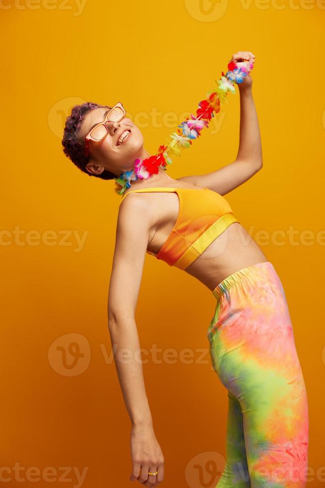 vrouw met bloemen hawaiiaans krans in de omgeving van haar nek heeft pret dansen en glimlachen in helder kleren Aan oranje achtergrond met zonnebril, levensstijl partij in hawaiiaans stijl foto