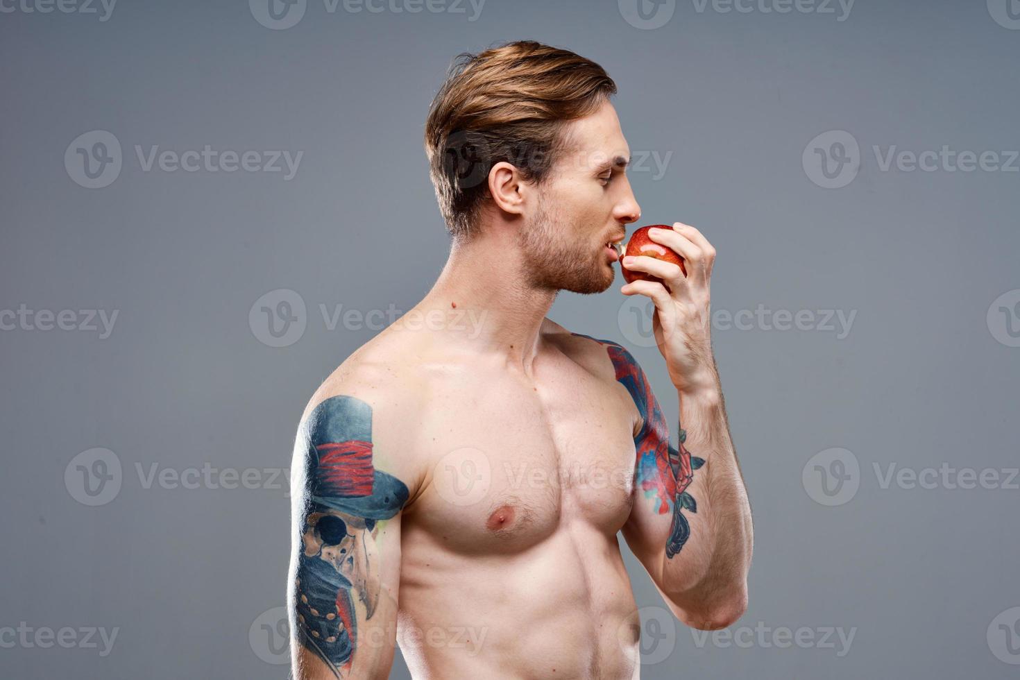 sexy atleet met gemotiveerd omhoog spieren eet een appel Aan een grijs achtergrond en een tatoeëren Aan zijn arm foto