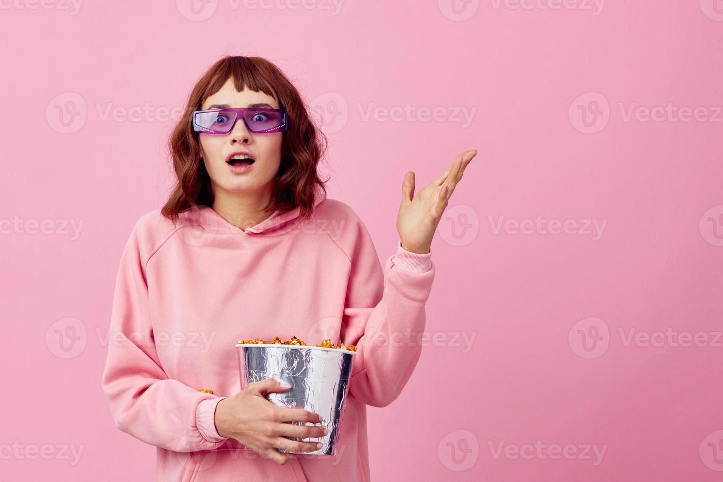 opgewonden dolblij gelukkig schattig roodharige dame in roze capuchon zonnebril met popcorn poseren geïsoleerd Aan over- roze studio achtergrond tonen kopiëren ruimte spandoek. mode bioscoop concept. vermaak aanbod foto