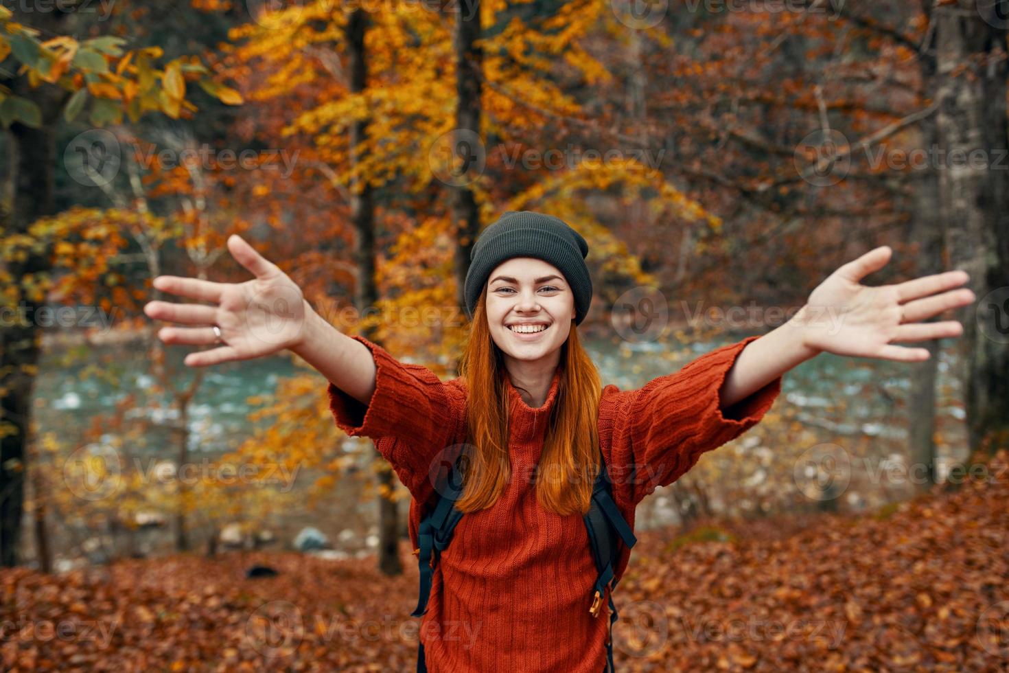 gelukkig vrouw met een rugzak reist in de park in herfst en gebaren met de handen van de rivier- in de achtergrond foto