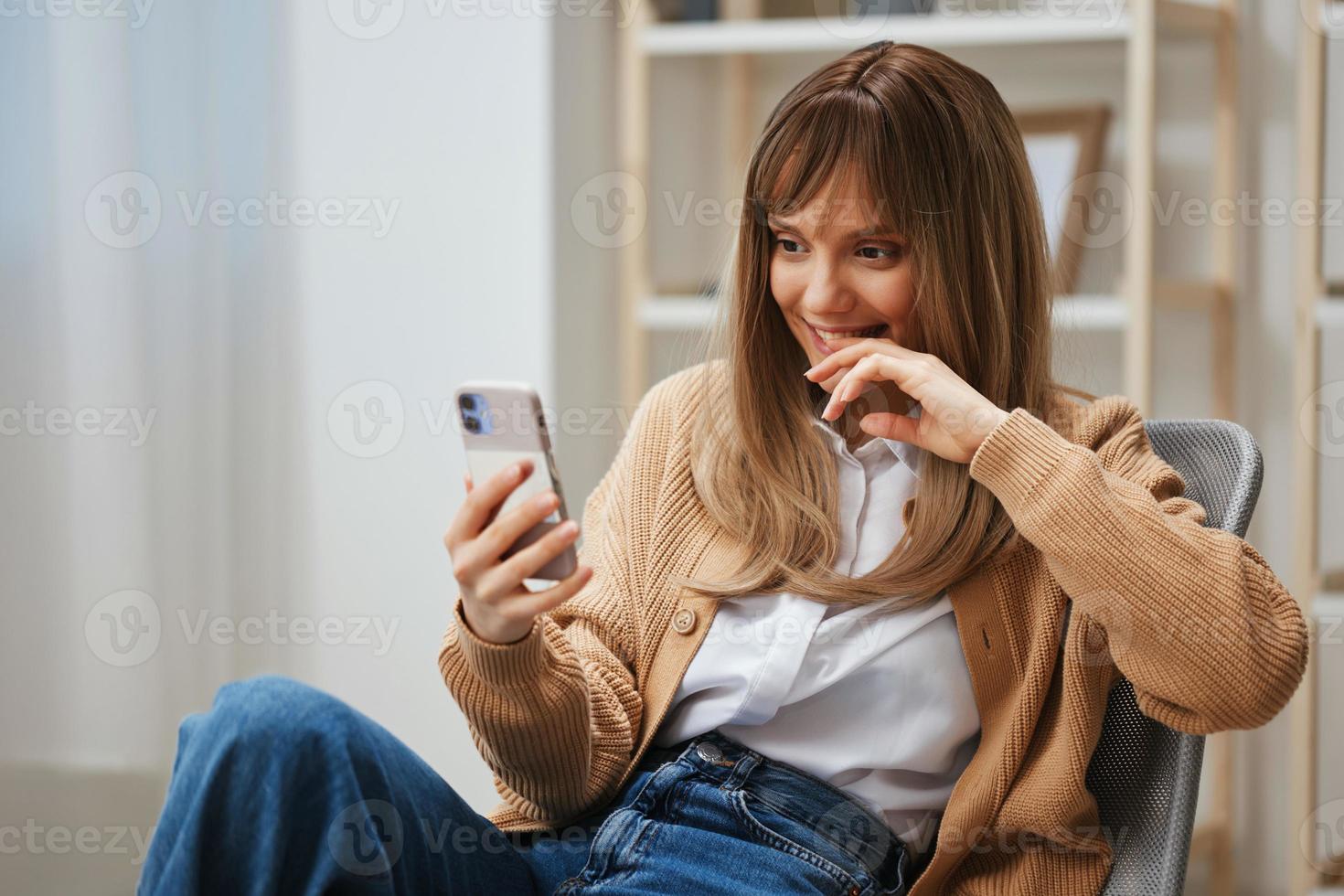 schattig jong blond dame in warm trui aan het doen selfie gebruik telefoon houden hand- in de buurt gezicht zittend in fauteuil Bij modern huis. pauze van werk, nemen een pauze, sociaal media in vrij tijd concept. kopiëren ruimte foto