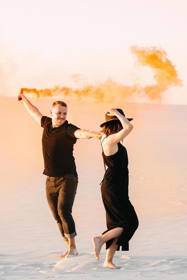 jongen en een meisje in zwarte kleren knuffelen en rennen op het witte zand met oranje rook foto