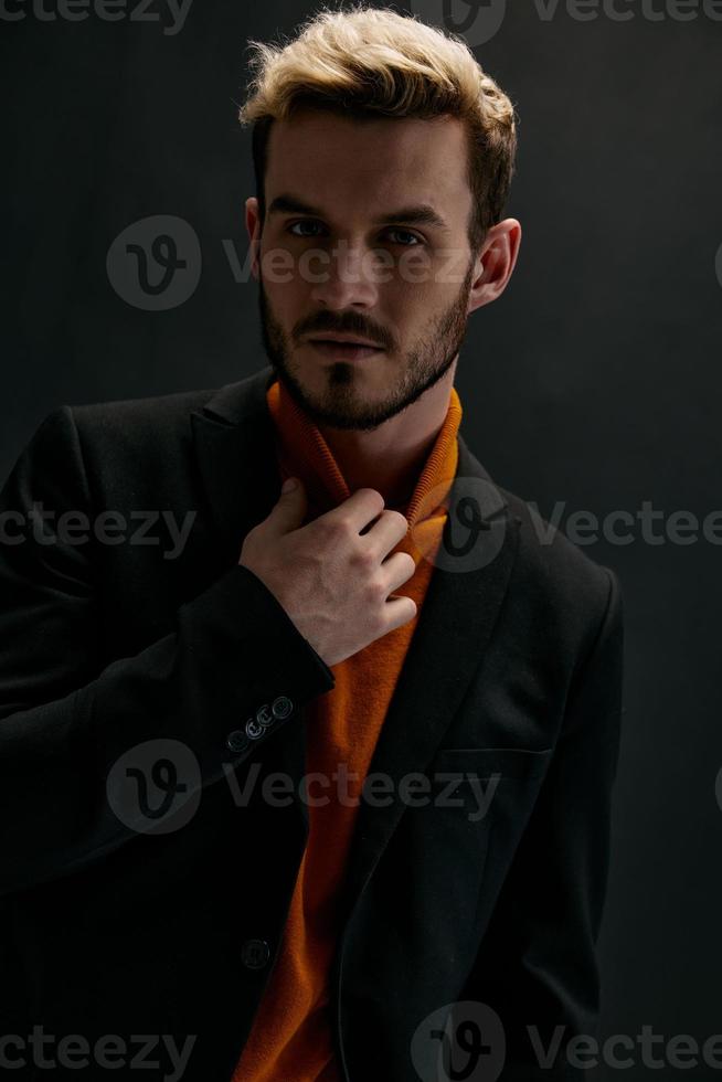 portret van een aantrekkelijk blond Mens Aan een donker achtergrond in een oranje trui en jas foto