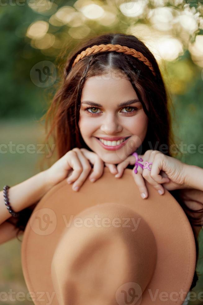 jong hippie vrouw Holding een hoed en glimlachen vriendelijke groet terwijl op zoek Bij de camera in natuur in de vallen foto