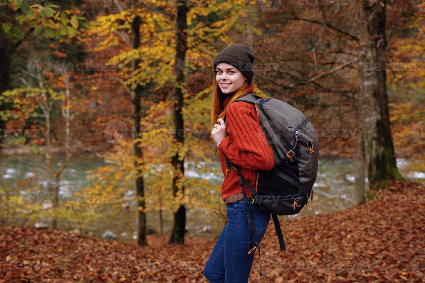 vrouw in herfst park met gedaald bladeren en rugzak Aan haar terug rivier- in de achtergrond foto