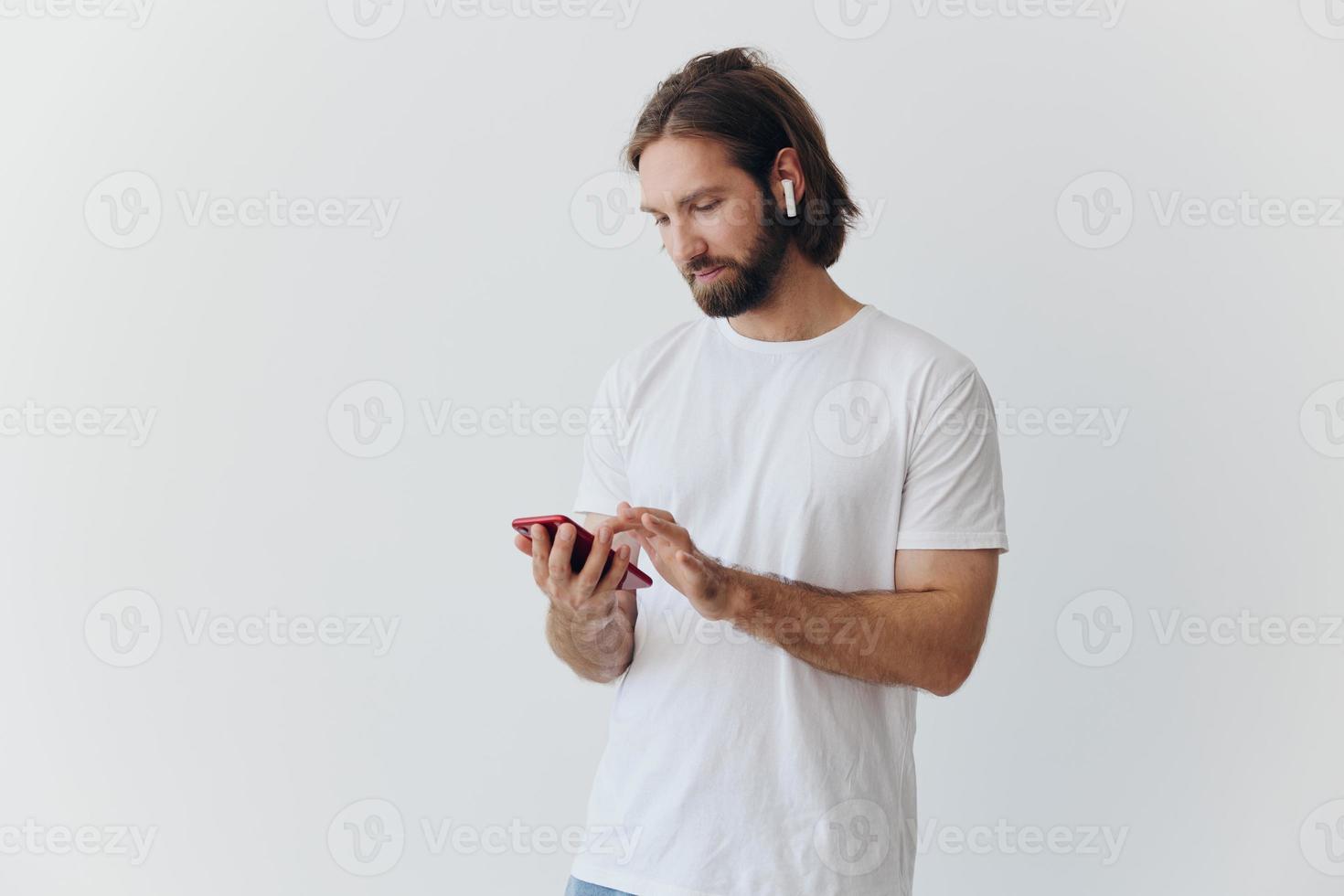 een Mens met een baard en lang haar- in een wit t-shirt en blauw jeans looks Bij zijn telefoon flippen door een online sociaal media voeden met hoofdtelefoons in zijn oren tegen een wit achtergrond foto