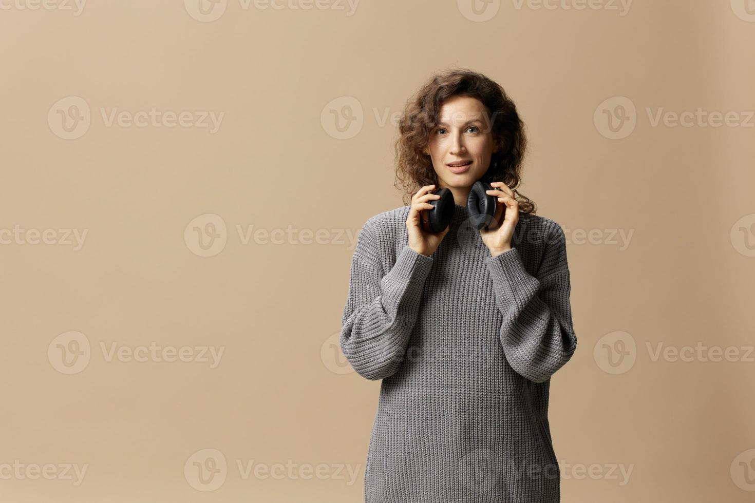 vrolijk genoten mooi gekruld mooi vrouw in grijs gewoontjes trui slijtage hoofdtelefoons looks Bij camera poseren geïsoleerd Aan over- beige pastel achtergrond. geluid streaming platform concept. kopiëren ruimte foto