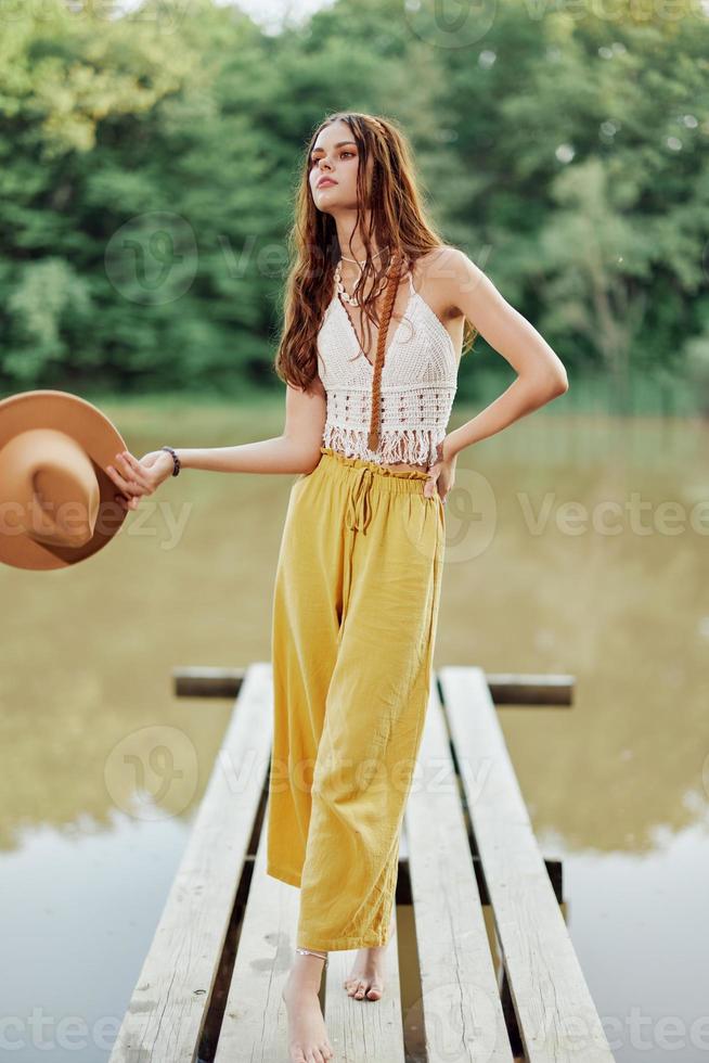 een jong vrouw in een hippie kijken en eco-jurk dansen buitenshuis door de meer vervelend een hoed en geel broek in de zomer zonsondergang foto