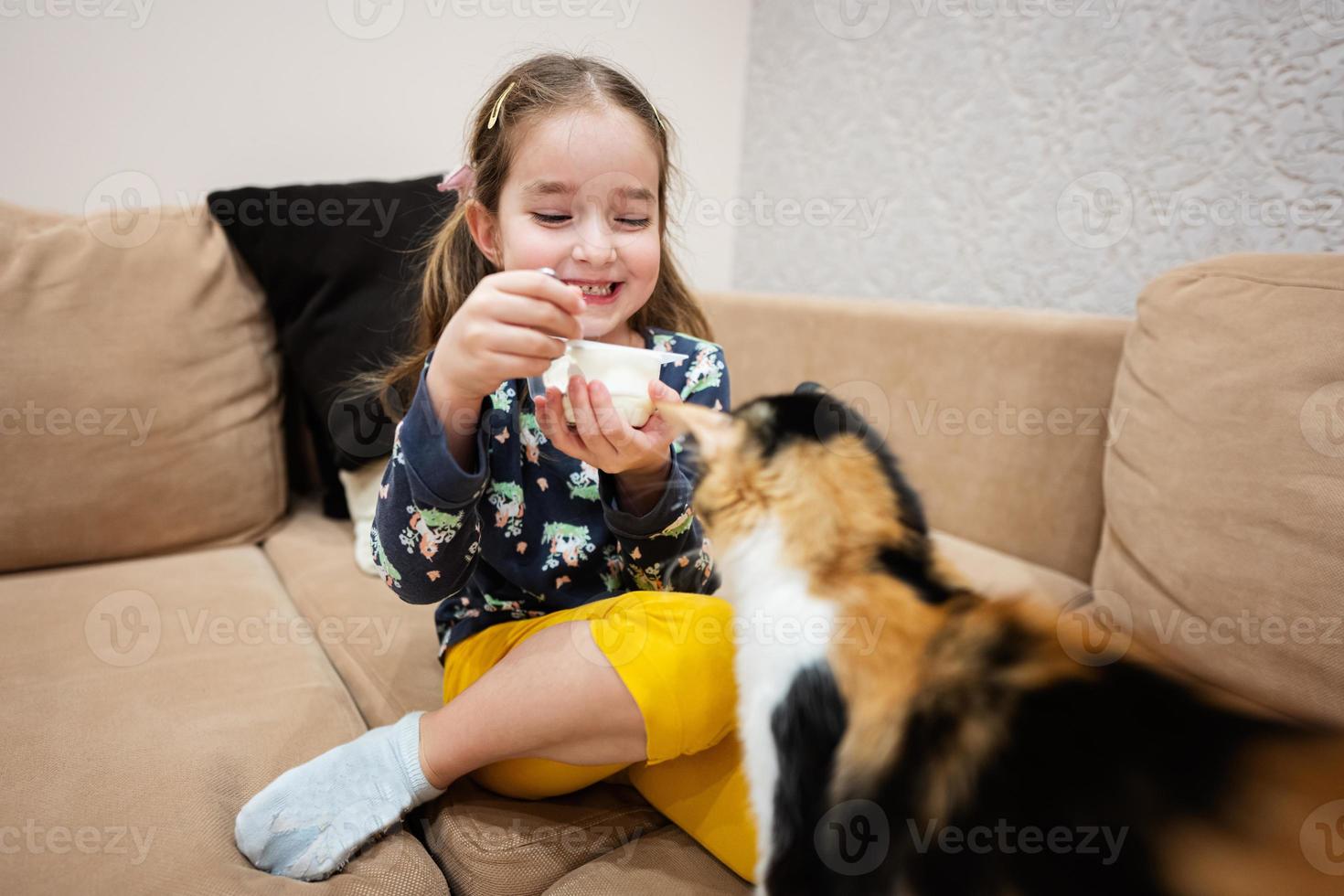 weinig meisje voeden de kat met yoghurt van een lepel Bij huis. foto