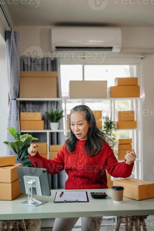 senior bedrijf eigenaar vrouw bereiden pakket doos en staand controleren online bestellingen voor leveren naar klant Aan tablet, laptop boodschappen doen online concept. Bij huis kantoor foto