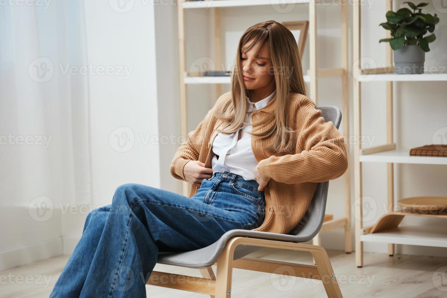 schattig gelukkig jong blond leerling dame freelancer in warm trui tintje taille zittend in fauteuil Bij modern huis interieur. pauze van werk, nemen een pauze, reflecties in vrij tijd concept. kopiëren ruimte foto