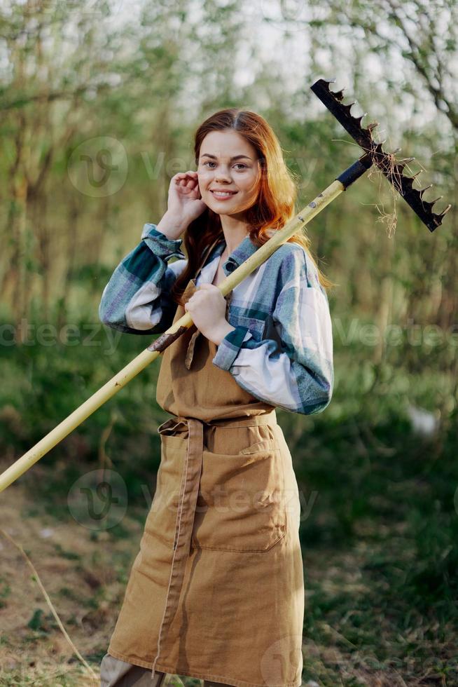 vrouw prachtig glimlachen boer in werk kleren en schort werken buitenshuis in natuur en Holding een hark naar verzamelen gras foto