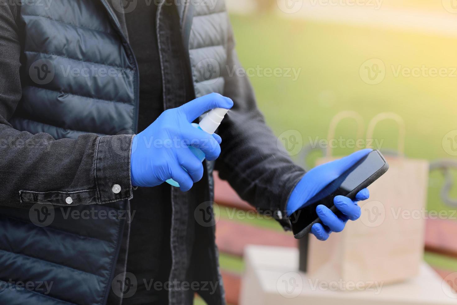 bezorgdienst koerier tijdens het coronavirus, covid-19 pandemie, koerier handen in handschoenen sproeien alcohol desinfecterende spray op een mobiel in de buurt van kartonnen dozen buitenshuis foto