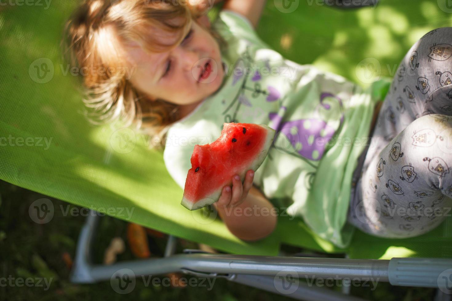 schattig weinig meisje aan het eten watermeloen Aan een dek stoel in de tuin in zomertijd foto