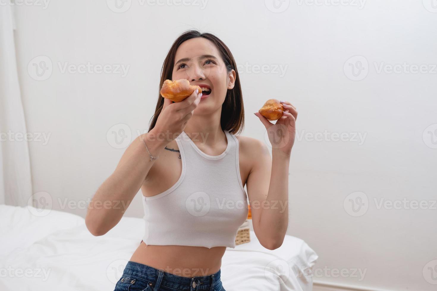 portret van wit Aziatisch vrouw met tatoeëren zittend Aan bed Holding brood en aan het eten rommel voedsel. zonder nemen zorg van Gezondheid tot het maakt u dik. dik concept. foto