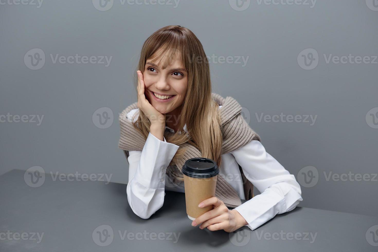 glimlachen dromen jong blond leerling dame freelancer in warm trui met meenemen koffie achterover leunen Aan hand- looks omhoog in grijs modern huis kantoor. koffie breken minnaar concept. kopiëren ruimte. koel aanbod foto