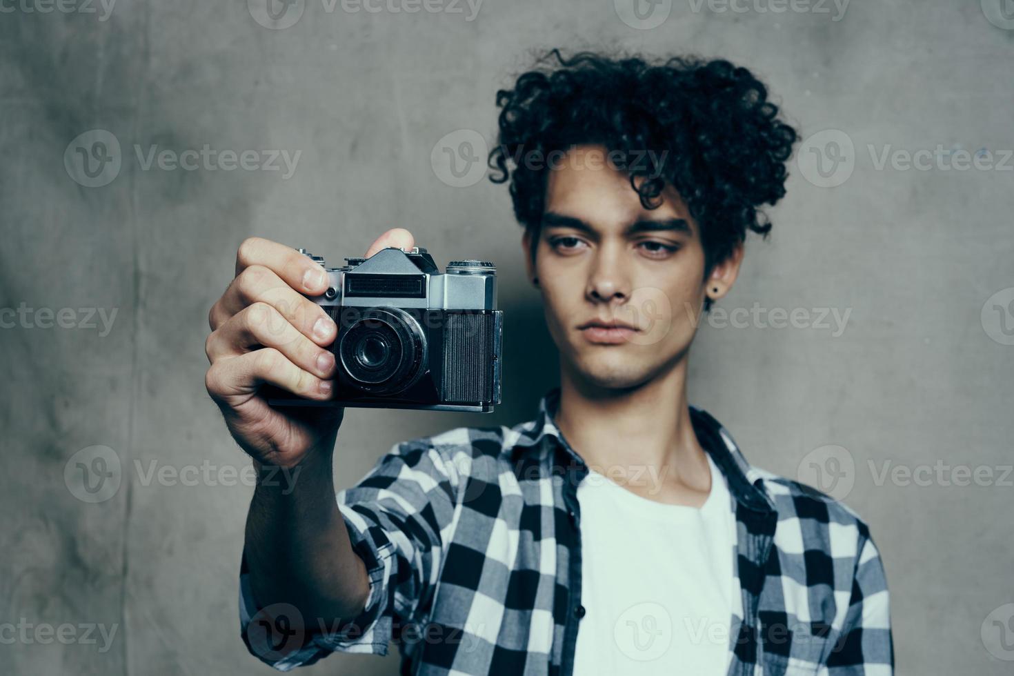 jong Mens met een camera in hand- en in een plaid t-shirt Aan een grijs achtergrond binnenshuis fotograaf foto