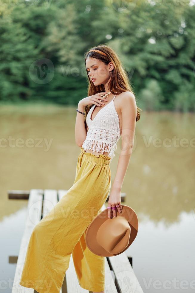 een jong vrouw glimlachen in een beeld van een hippie en eco-jurk dansen in natuur door de meer vervelend een hoed en geel broek in de zomer zonsondergang foto