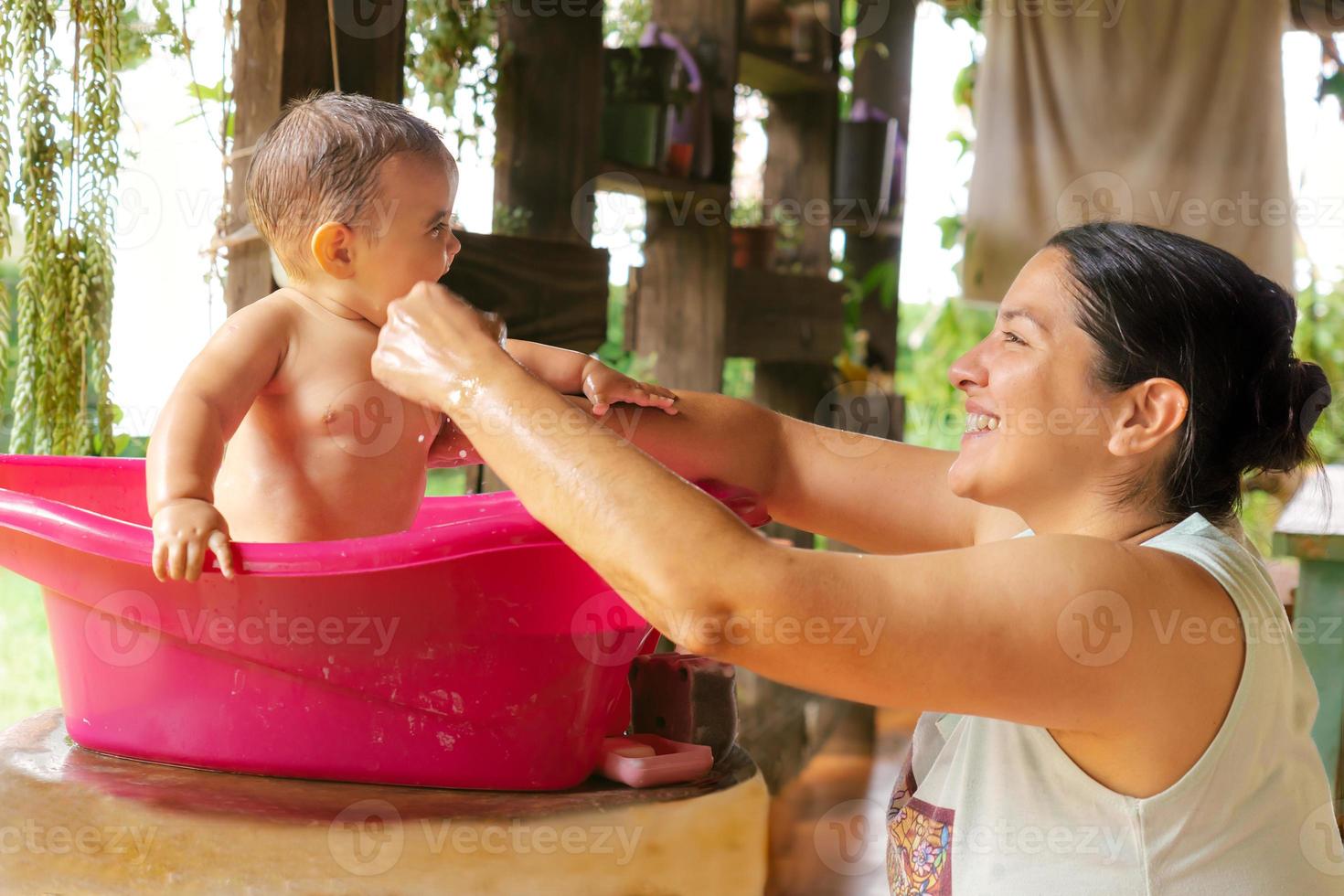 een moeder liefdevol baadt haar baby meisje in een emmer buitenshuis, het verstrekken van inschrijving zorg en bemoedigend mooi zo hygiëne, terwijl ze binding in de warm zonneschijn. foto