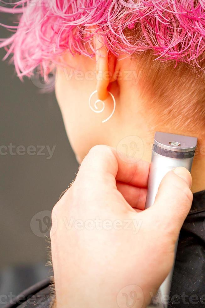 terug visie van kapper hand- scheren nek en nek met elektrisch trimmer van jong Kaukasisch vrouw met kort roze haar- in schoonheid salon. foto