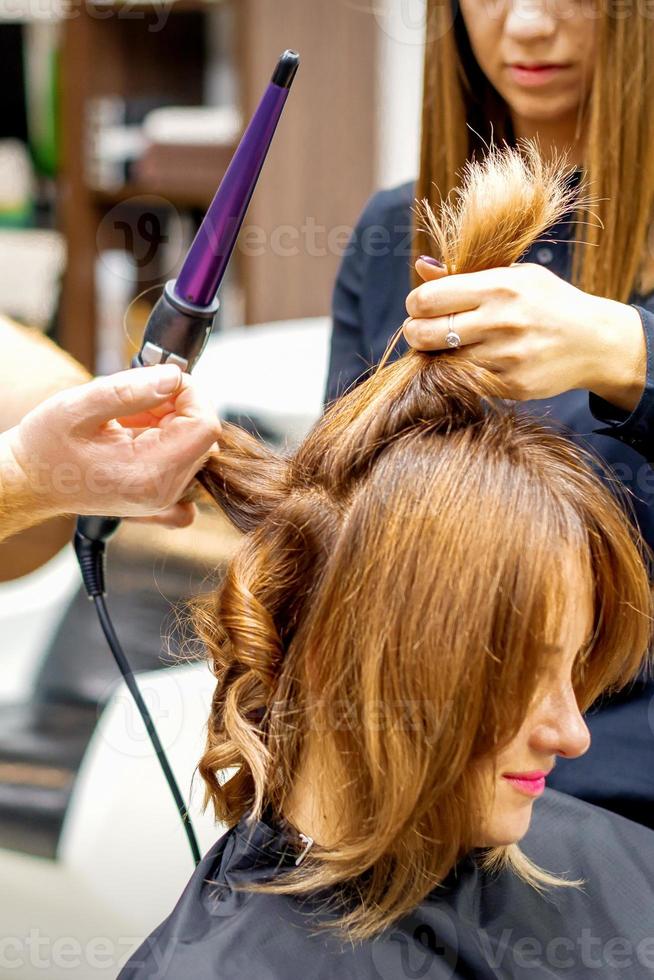 twee Haarstylisten gebruik makend van curling ijzer Aan klanten lang bruin haar- in een schoonheid salon. foto