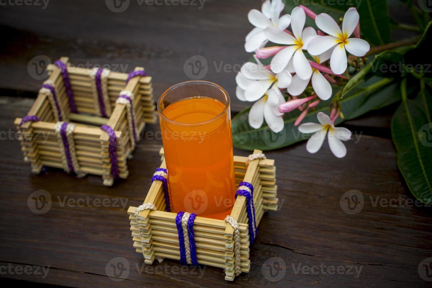 een glas van oranje sap met water glas houder gemaakt van bamboe stokjes en vezel. decoratief houten tafel met mooi plumeria bloemen. bamboe glas houder. foto