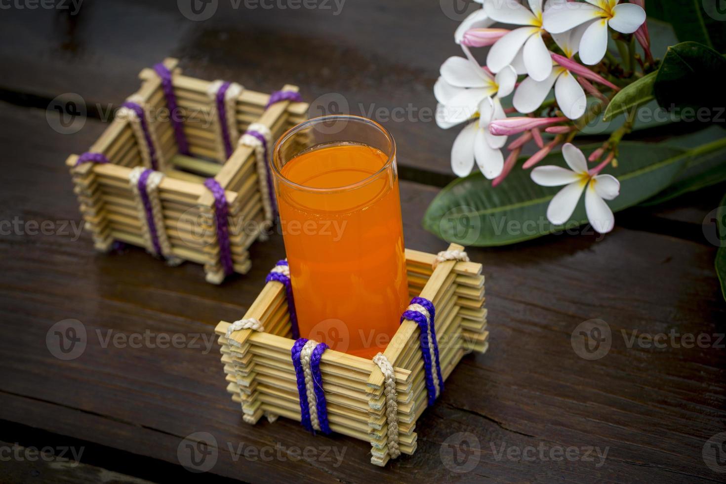 een glas van oranje sap met water glas houder gemaakt van bamboe stokjes en vezel. decoratief houten tafel met mooi plumeria bloemen. bamboe glas houder. foto