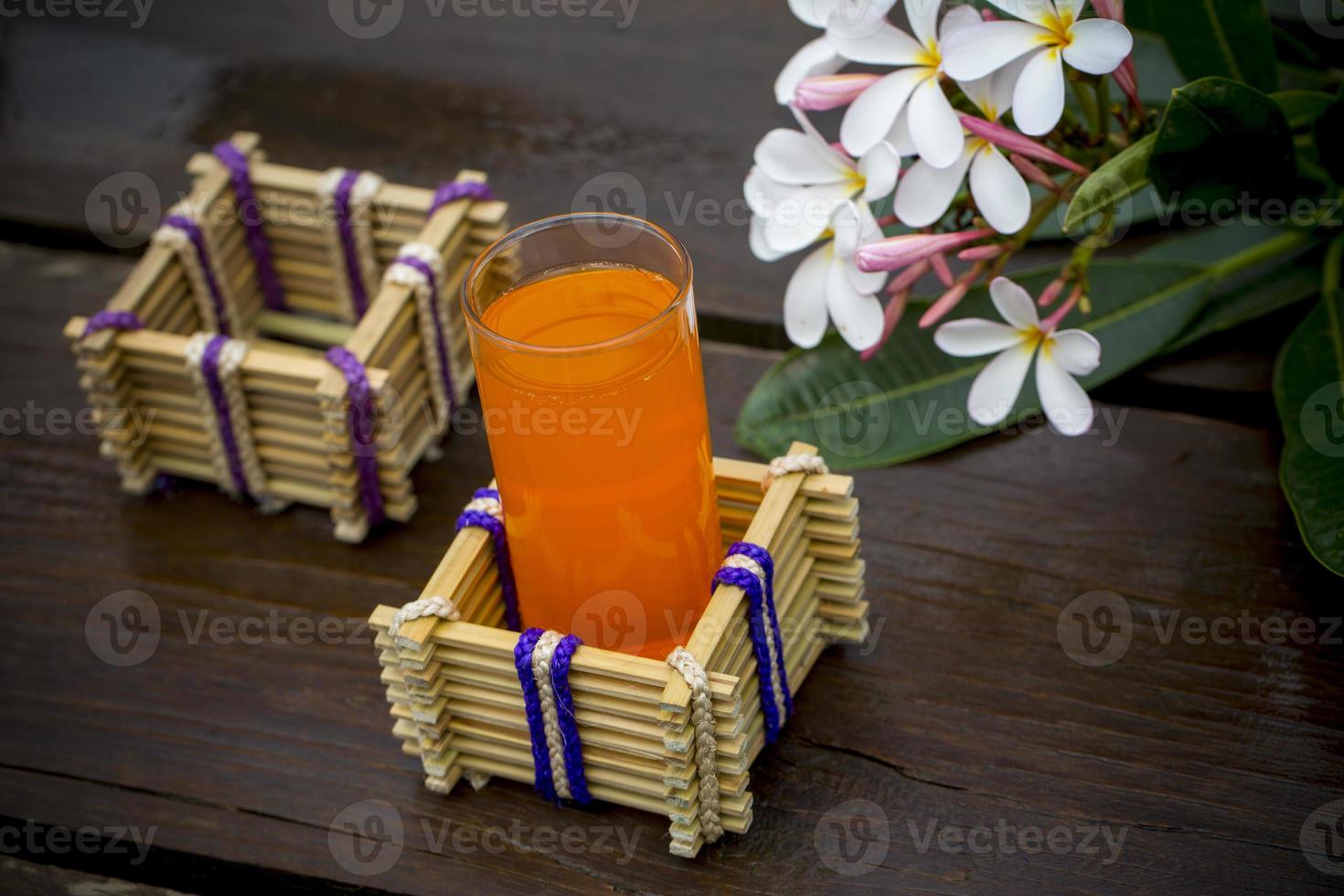 een glas van oranje sap met water glas houder gemaakt van bamboe stokjes en vezel. decoratief houten tafel met mooi plumeria bloemen. bamboe glas houder. foto