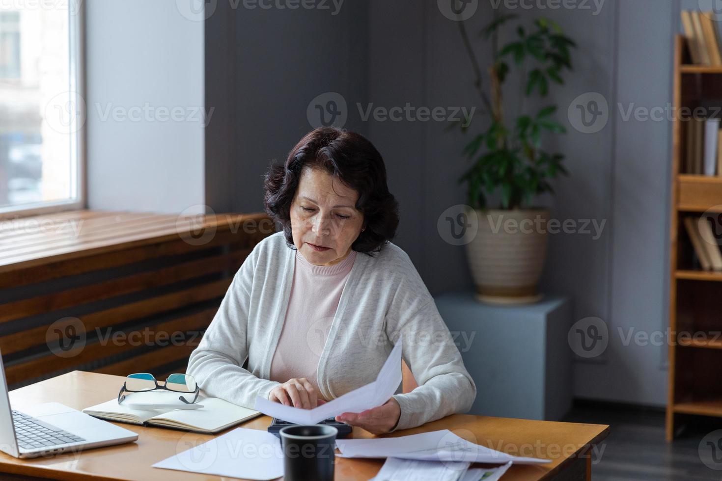 midden- oud senior vrouw zitten met laptop en papier document, nadenkend ouder volwassen dame lezing papier Bill betalen online Bij huis beheren bank financiën berekenen belastingen planning lening schuld pensioen betaling foto