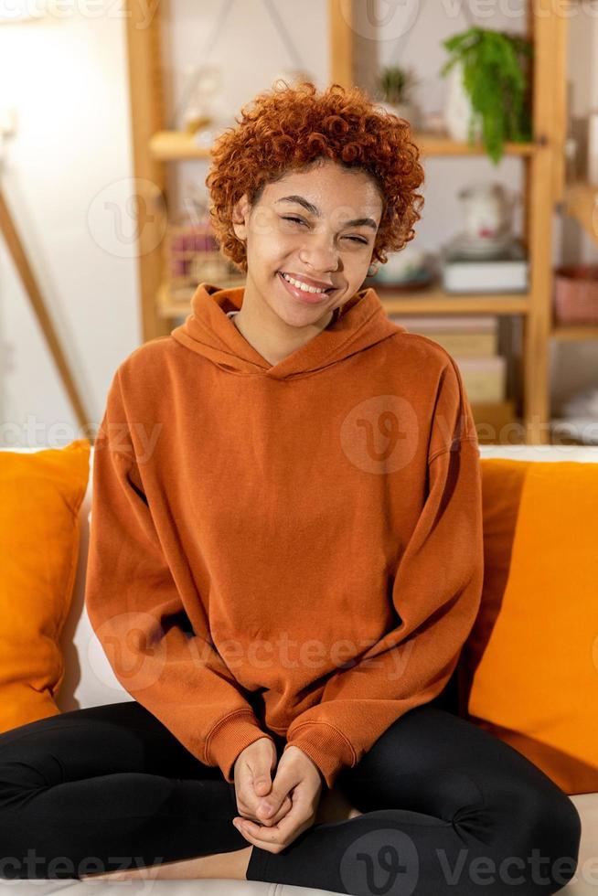 mooi Afrikaanse Amerikaans meisje met afro kapsel glimlachen zittend Aan sofa Bij huis binnen. jong Afrikaanse vrouw met gekruld haar- lachend. vrijheid geluk zorgeloos gelukkig mensen concept. foto