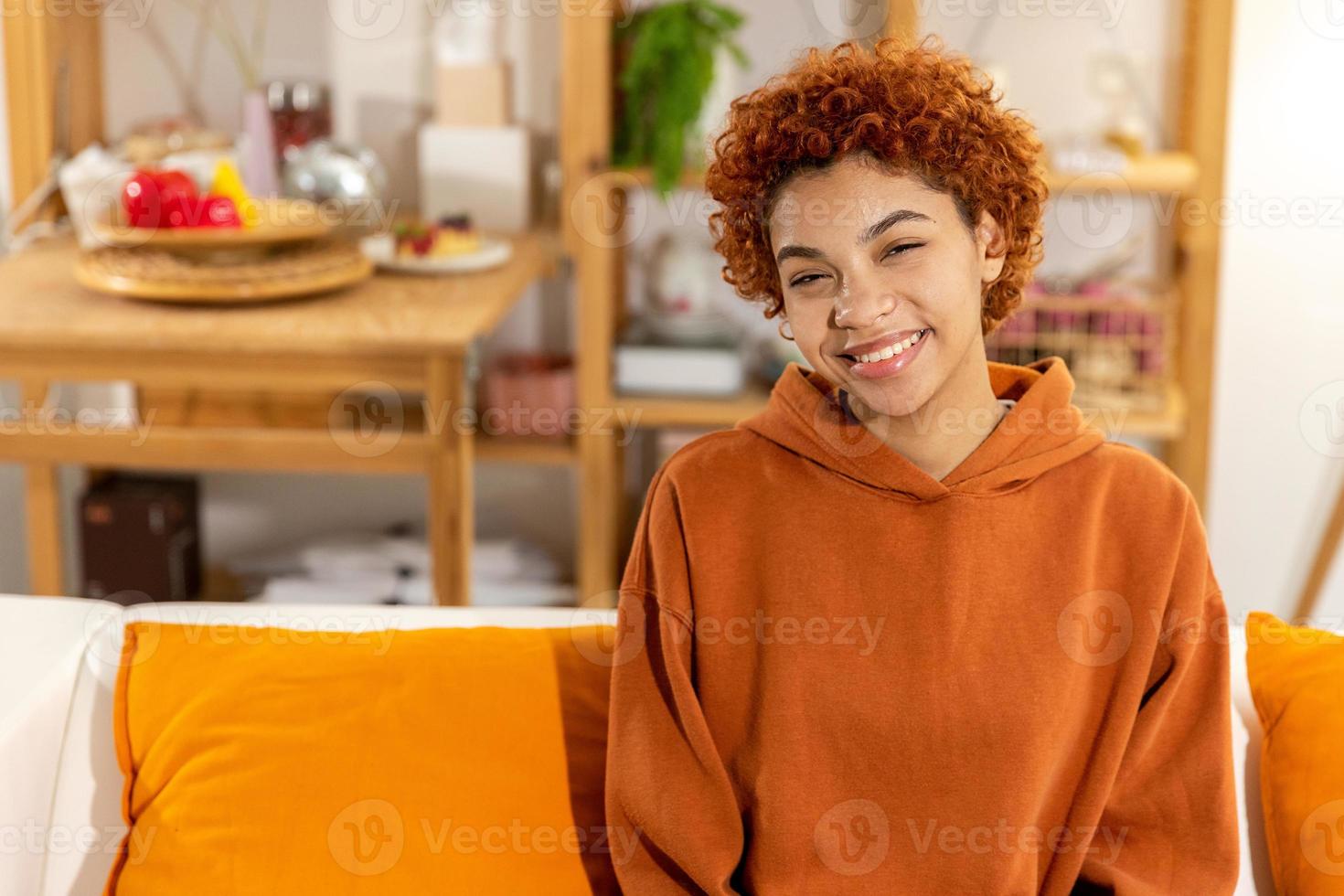 mooi Afrikaanse Amerikaans meisje met afro kapsel glimlachen zittend Aan sofa Bij huis binnen. jong Afrikaanse vrouw met gekruld haar- lachend. vrijheid geluk zorgeloos gelukkig mensen concept. foto