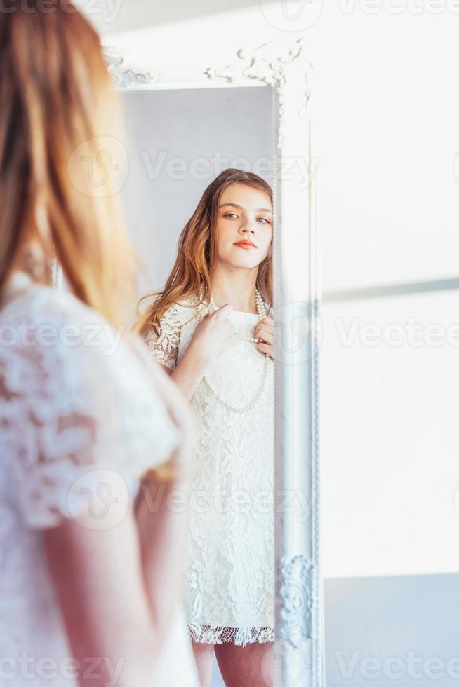 schoonheid bedenken ochtend- rutine liefde jezelf concept. jong tiener- meisje op zoek Bij reflectie in spiegel. jong positief vrouw vervelend wit jurk poseren in helder licht kamer tegen wit muur. foto