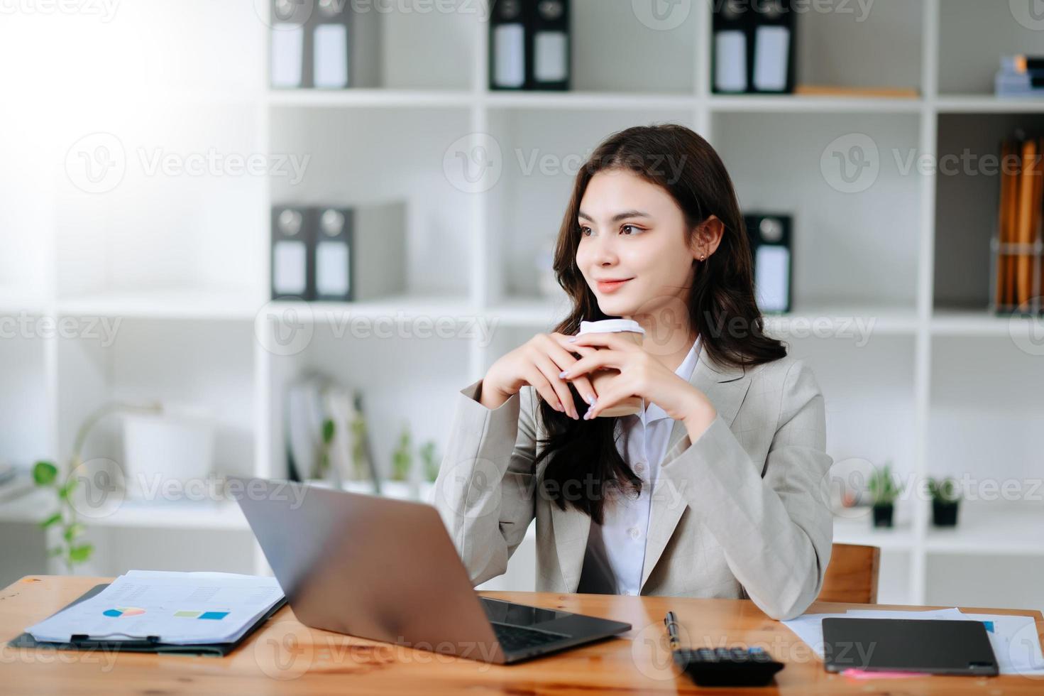 zelfverzekerd mooi Aziatisch zakenvrouw typen laptop computer en digitaal tablet terwijl Holding koffie Bij kantoor foto