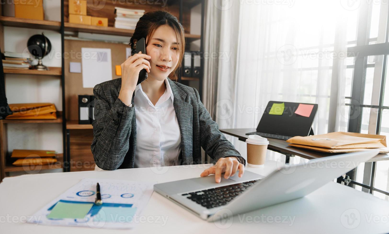 bedrijf Aziatisch vrouw pratend Aan de telefoon en gebruik makend van een laptop met een glimlach terwijl zittend Bij kantoor foto