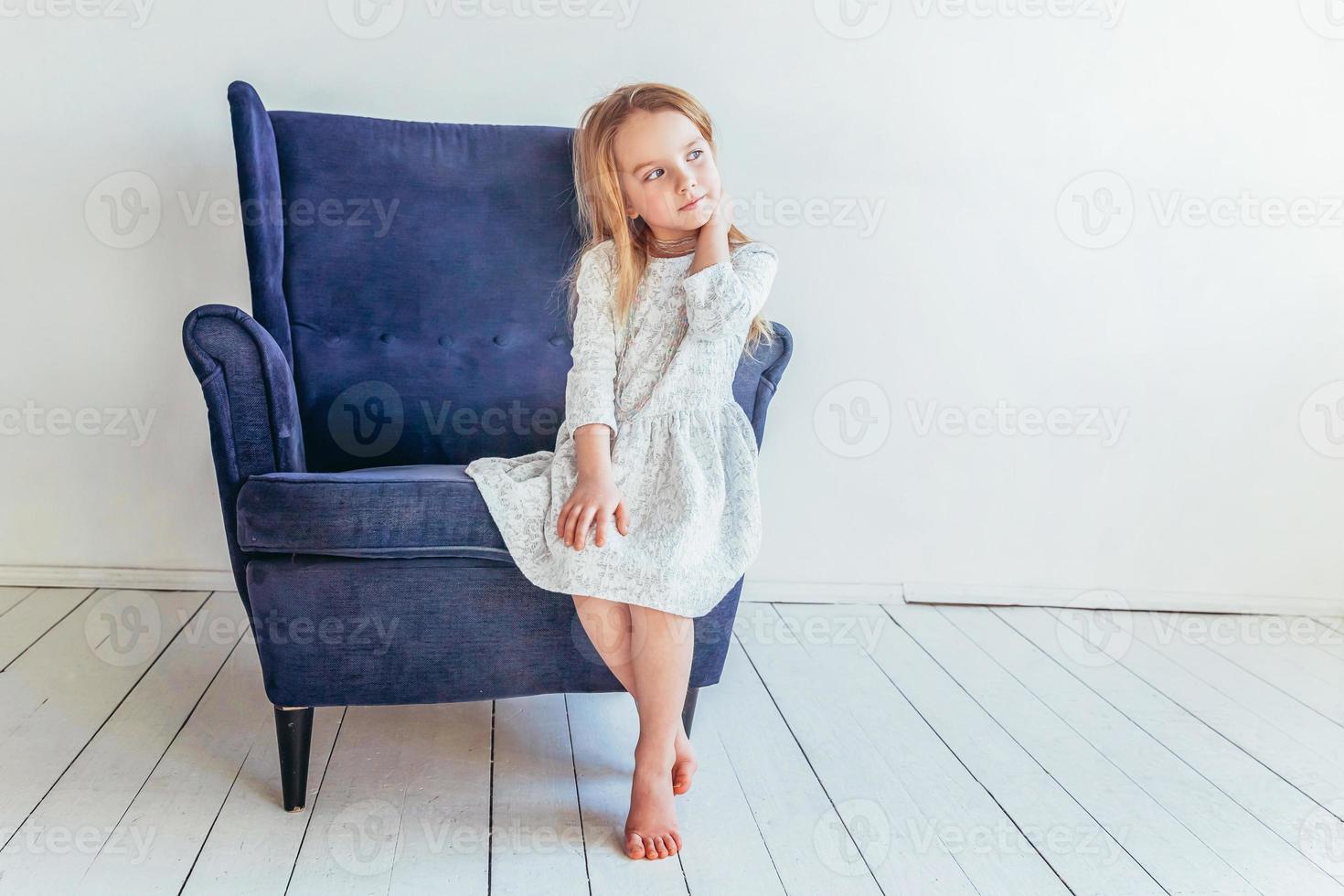 zoet weinig meisje in wit jurk zittend Aan modern knus blauw stoel ontspannende in wit helder leven kamer Bij huis binnenshuis. kinderjaren schoolkinderen jeugd kom tot rust concept. foto