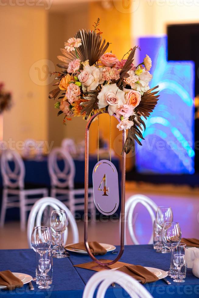 feestelijk tafel met blauw lap, servetten, platen, bestek en bril bereid voor bruiloft gasten in restaurant. mooi bloemen arrangement in goud metaal vaas met aantal vier. verbazingwekkend porties foto