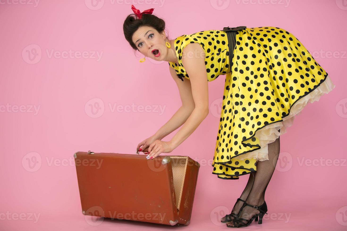 mooi vastpinnen meisje opent een retro koffer. retro vrouw in geel polka punt jurk. de emotie van verrassing. foto